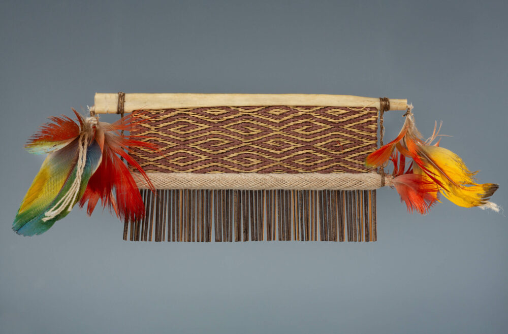 Objekt aus Brasilien mit rot-gelb gewebtem Stoffmuster und an den Seiten bunten Vogelfedern.
