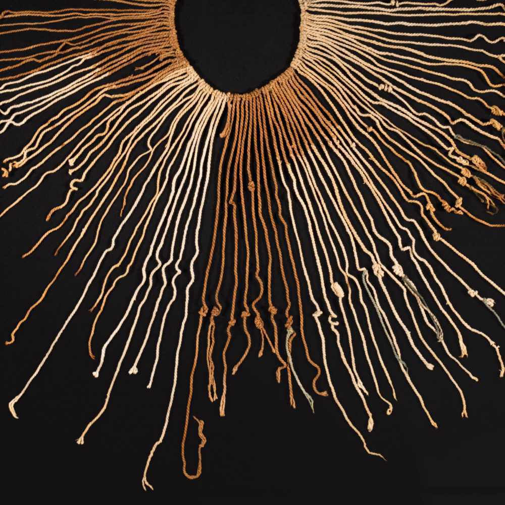 Im Halbkreis angeordnetes Knotenschnurbündel vor schwarzem Hintergrund. Die Schnüre sind beige, braun und grün.