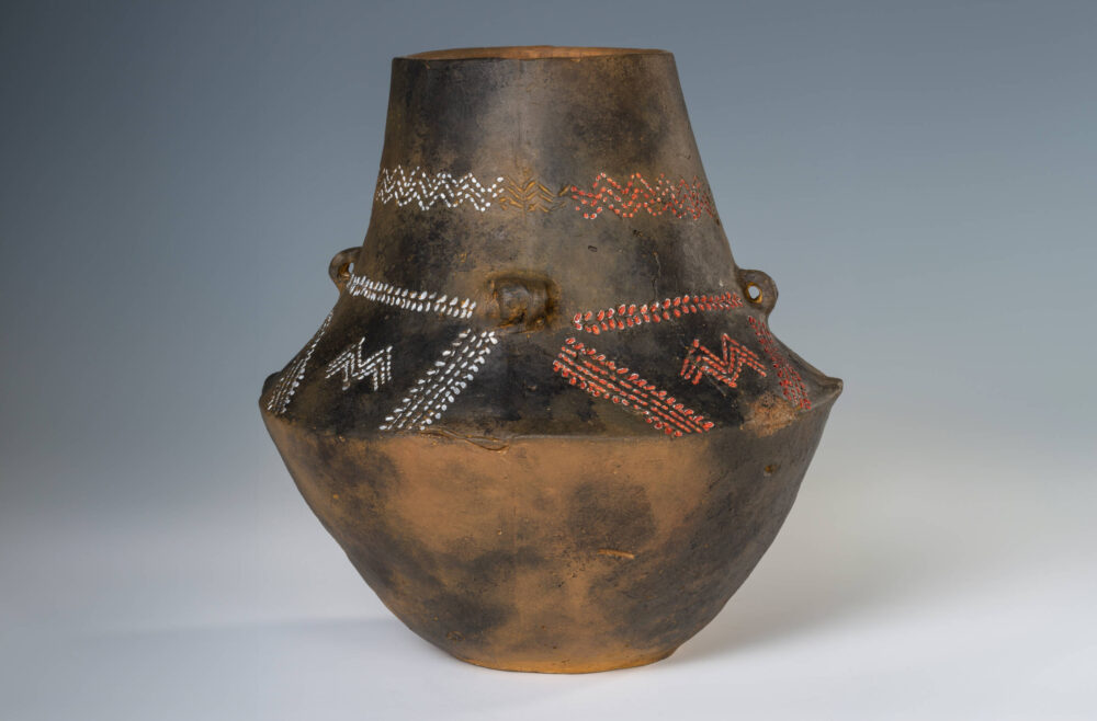 Bauchiges, dunkelbraunes Tongefäß mit Punkt- und Strich-Verzierungen in weiß und rot.
