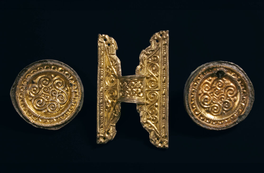 Zwei goldene, runde Scheibenfibeln rahmen eine ebenfalls goldene gleicharmige Bügelfibel. Sie tragen alle ein Spiralmuster.