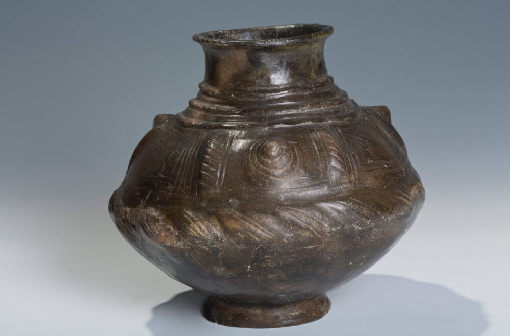 Ein dunkelbraunes Keramikgefäß mit bauchiger Mitte und kleiner runder Öffnung oben, mit Verzierungen.