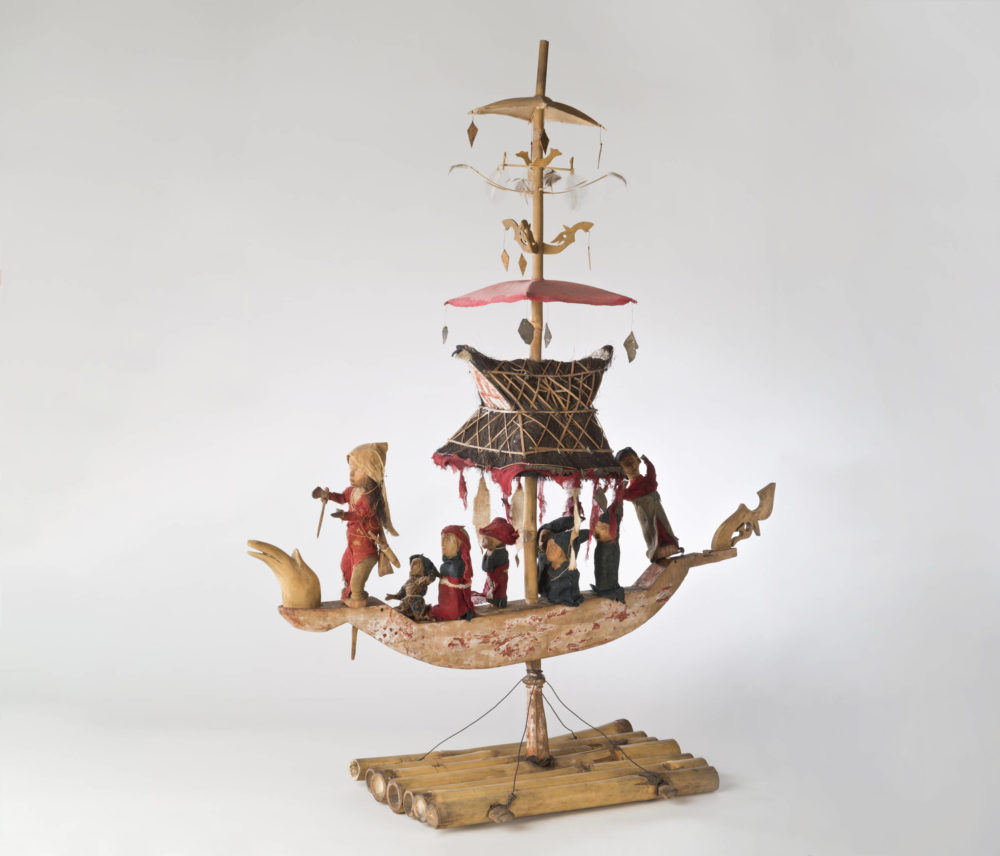 Modell eines indonesischen Seelenbootes, das mit hohem Mast und acht kleinen Modellfigürchen bestückt ist.
