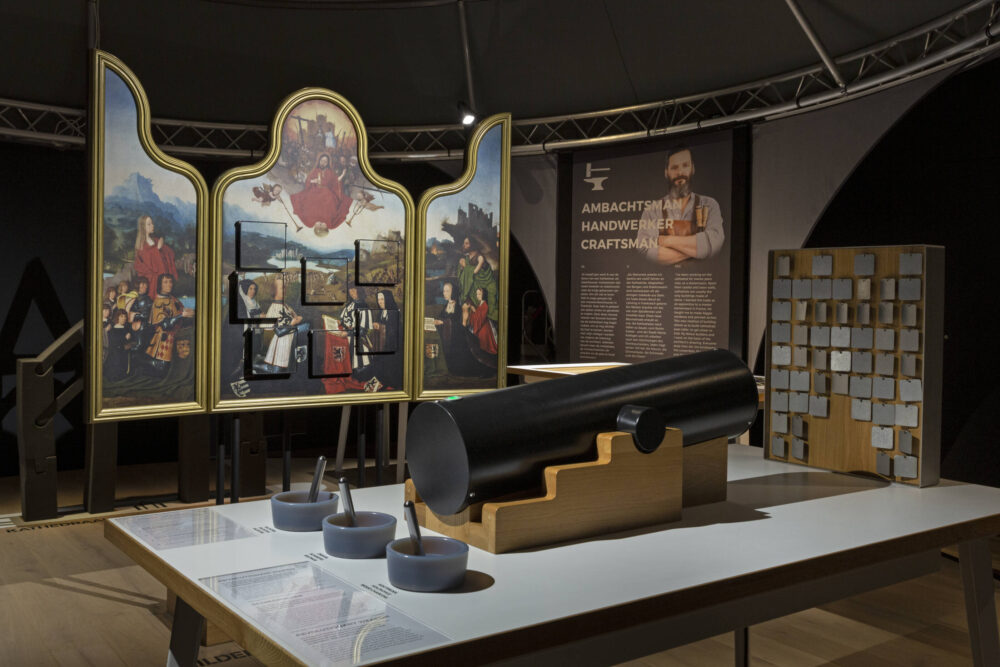 Blick in die Ausstellung, im Vordergrund eine Mitmachstation, die an eine Kanone erinnert, im Hintergrund eine Replik eines Altarretabels mit Klappen zum öffnen.