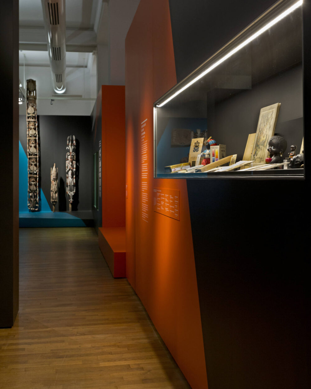 Ausstellungeinblick, Vitrine im Vordergrund auf der rechten Seite, große ethnologische Objekte im Hintergrund.