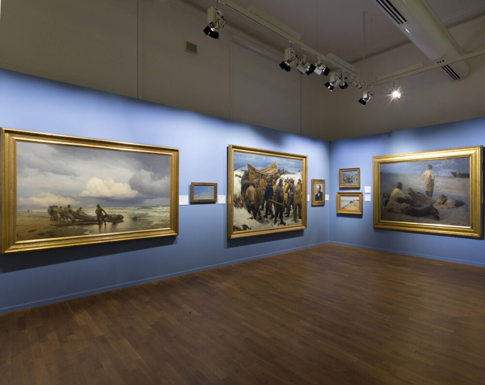 Blick in eine Kunstausstellung mit Gemälden verschiedener Größe in goldenen Rahmen.