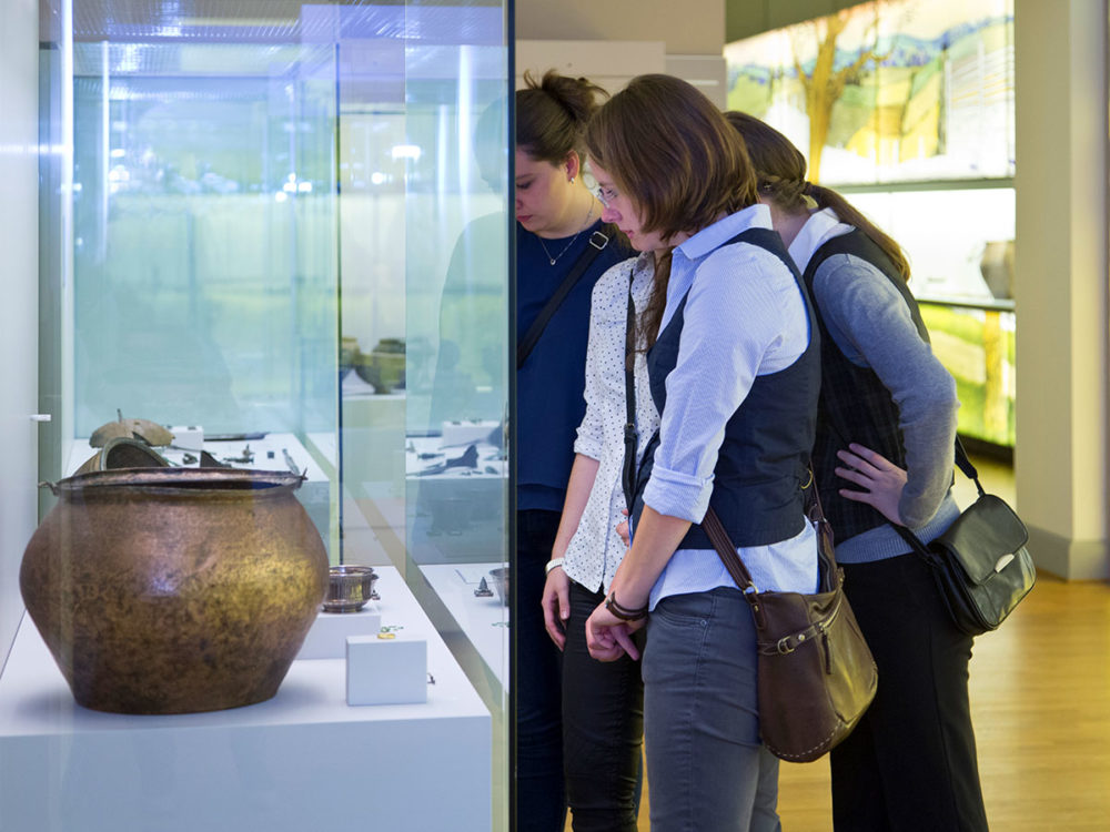 Drei junge Frauen schauen in eine Vitrine mit archäologischen Funden in den MenschenWelten des Landesmuseums.