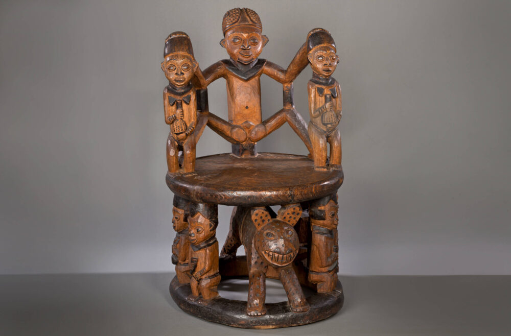 Verschiedene menschlichen Figuren und ein Tier tragen die Sitzplatte und bilden die Lehne eines hölzernen Throns.