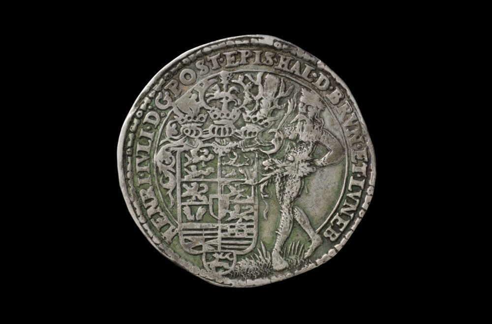 Leicht abgegriffene Münze mit großem Wappen und einer danebenstehenden Person sowie einer Umschrift.