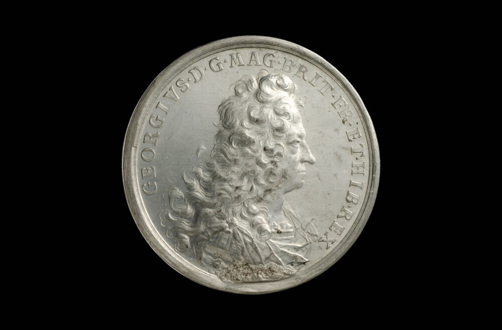 Silbern glänzende Medaille mit Georg I. als König von England im Profil und einer darauf hinweisenden Umschrift.