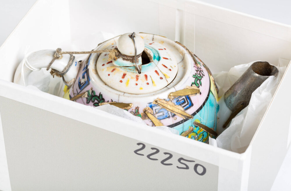 Blick in einen Karton, in dem eine sichtbar reparierte Porzellankanne mit bunter Bemalung aufbewahrt wird.