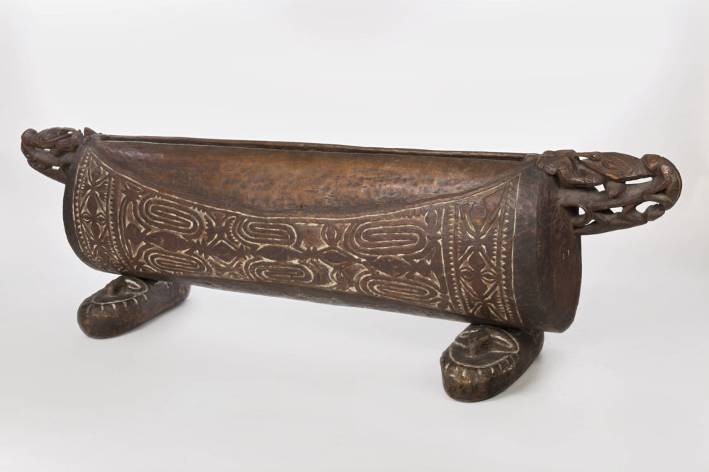 Aus dunkelbraunem Holz gefertigte Schlitztrommel mit Füßen, seitlichen Griffen und umfangreicher Schnitzverzierung.