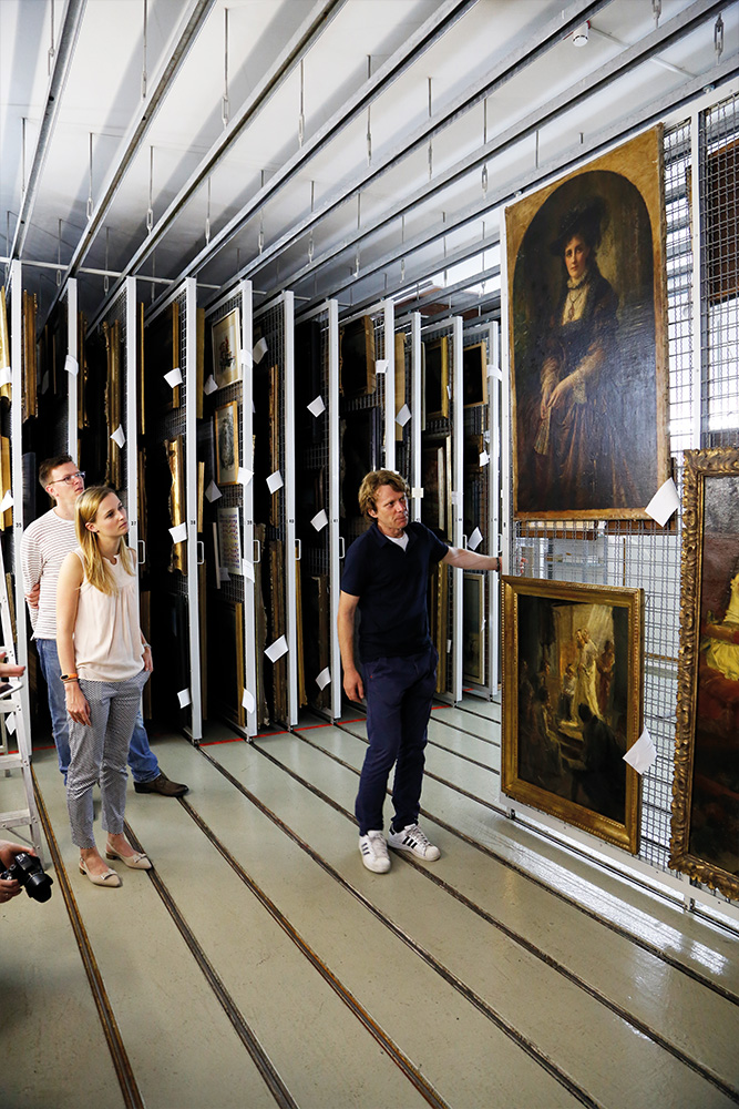 Blick ins Kunstdepot, in dem der Kurator zwei Besuchern Gemälde zeigt, die an einer Gitterschiebewand hängen.