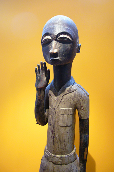 Afrikanische Holzfigur eines Menschen mit gräulicher Uniform, die wie zum Gruß die Hand hebt.